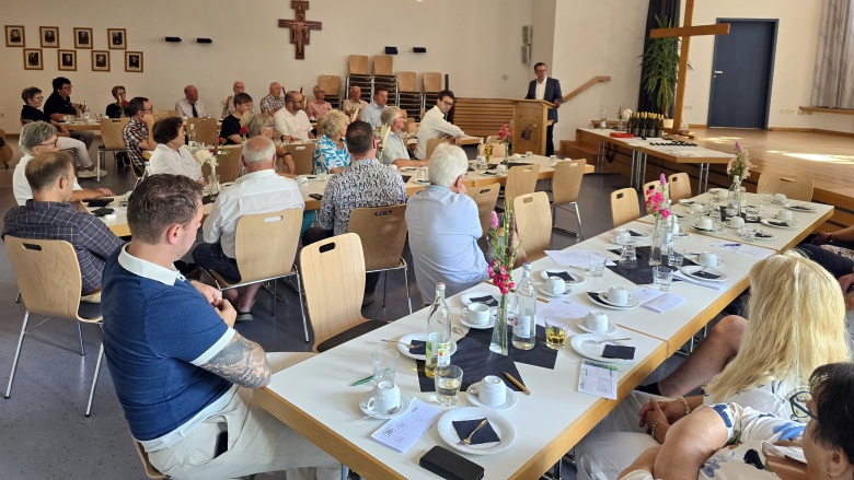 Saal mit mehreren für Kaffee und Kuchen gedeckten Tischen. An den Tischen sitzen ca. 25 Personen. An einem Rednerpult steht der Kreisvorsitzende der CDU Frederik Schmitt und spricht zu den Personen im Saal