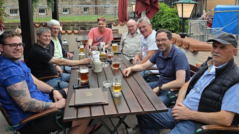 Acht männliche Personen sitzen an einem Tisch auf einer Terrasse. Auf dem Tisch Getränke.